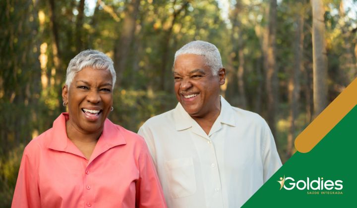 Um casal de idosos sorri alegremente enquanto estão juntos em um parque arborizado, ilustrando o post sobre "Bem-estar na terceira idade: como aproveitar melhor a vida"