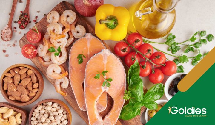 Variedade de ingredientes frescos, incluindo salmão, camarão, tomate cereja, pimentão amarelo, amêndoas, grão-de-bico, ervas e azeite de oliva em uma superfície leve, ilustrando o post "Como a dieta mediterrânea ajuda no envelhecimento saudável?"