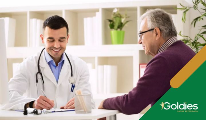Um médico de jaleco branco sorri enquanto faz anotações, sentado em uma mesa em frente a um homem mais velho usando óculos, ilustrando o post "Quando procurar um geriatra?"