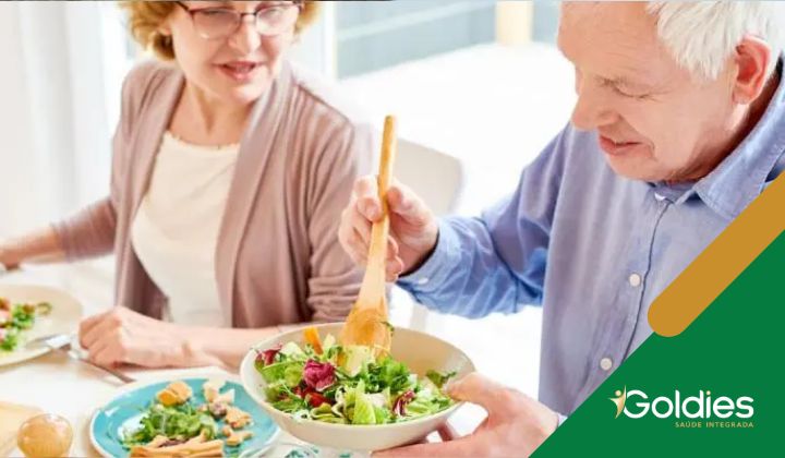 Um homem idoso serve salada de uma tigela enquanto está sentado à mesa de jantar com uma mulher idosa. Ambos estão sorrindo. A mesa está posta com comida, ilustrando o post "A importância das vitaminas na alimentação dos maduros"