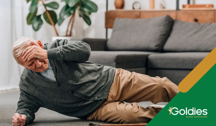 Um homem idoso está deitado no chão de uma sala de estar, parecendo ter caído. Ele está segurando a cabeça, parecendo angustiado, ilustrando o post "O que causa a fraqueza nas pernas dos maduros?"