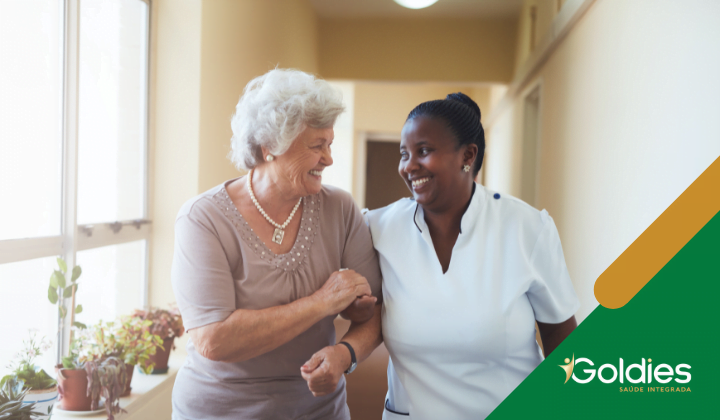 Mulher idosa e cuidadora caminhando juntas, sorrindo, em um corredor bem iluminado com plantas perto da janela, ilustrando o post "A rotina dos maduros necessita de cuidados"