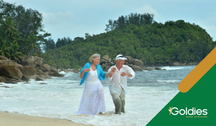 Casal de seniores na praia, felizes com as ondas batendo a seus pés, ilustrando o post "Cuidados com maduros no verão"