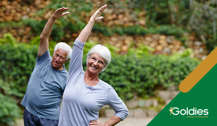 Homem e mulher idosos fazendo exercícios de alongamento ao ar livre em um jardim. Ambos estão vestindo roupas esportivas cinza e sorrindo, ilustrando o post "5 MANEIRAS DE TER UMA VIDA ATIVA NA TERCEIRA IDADE"