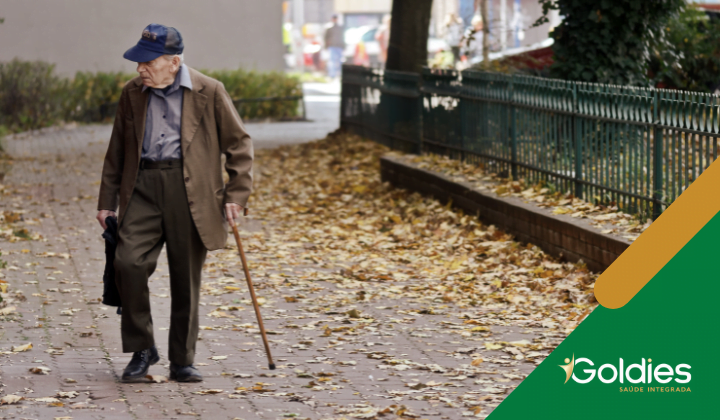 Homem idoso com casaco marrom e boné azul caminha com uma bengala em um caminho coberto de folhas ao lado de uma cerca verde, ilustrando o post "Caminhada para os seniores: por que apostar nessa atividade?"