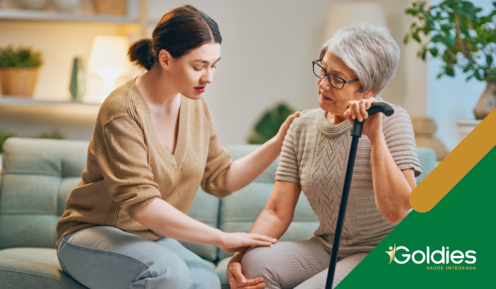 Uma jovem auxilia uma idosa de cabelos curtos e grisalhos, sentada em um sofá e segurando uma bengala, ilustrando o post "Quais cuidados devemos ter com a saúde do maduro?"