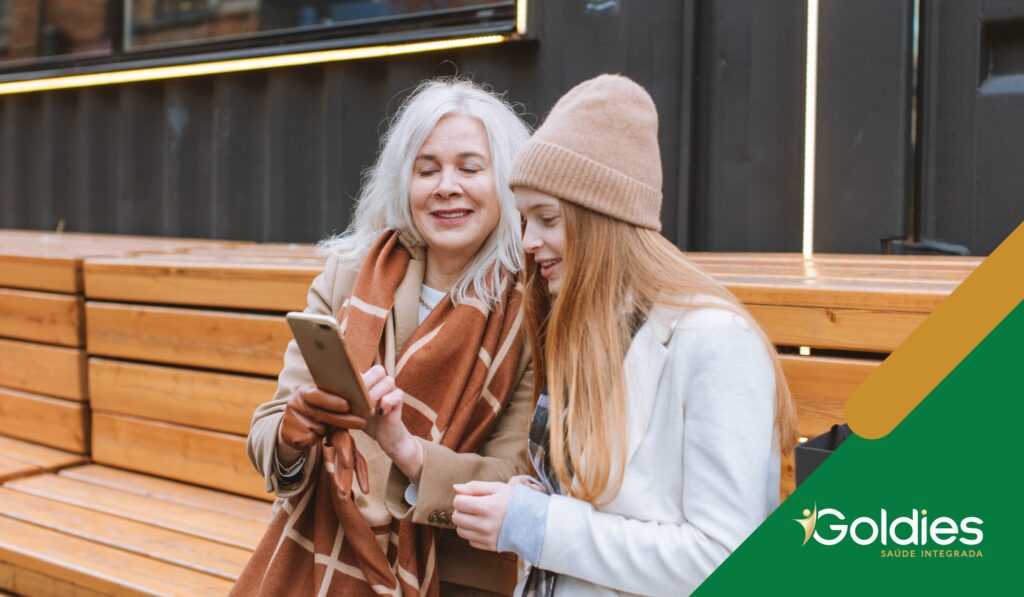 Mãe e filha, sorrindo em um momento de descontração, olhando para um celular e utilizando juntas um aplicativo, ilustrando o post "Aplicativos de saúde e bem-estar: qual a sua importância?"