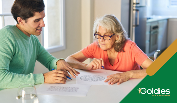 Um homem de suéter verde e uma mulher idosa de óculos sentam-se à mesa, envolvidos em uma atividade criativa ou terapêutica envolvendo um padrão detalhado no papel. O cenário é uma sala iluminada, ilustrando postagem relacionada ao Mal de Alzheimer