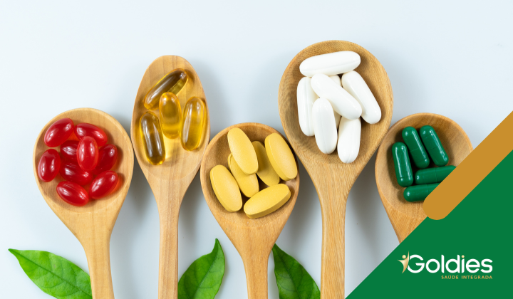 Cinco colheres de madeira seguram pílulas e cápsulas de cores diferentes: vermelho, amarelo, branco, verde e translúcido. Folhas verdes são colocadas sob as colheres. Um design verde e dourado com o texto Goldies está no canto inferior direito.