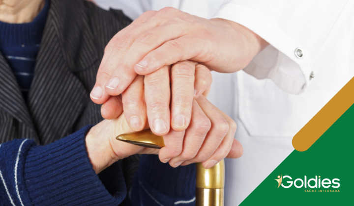 Um médico gentilmente coloca suas mãos sobre as mãos de uma pessoa idosa apoiadas em uma bengala. O médico veste um jaleco branco, e a pessoa idosa veste um suéter azul escuro. O logotipo do Goldies está no canto inferior direito.