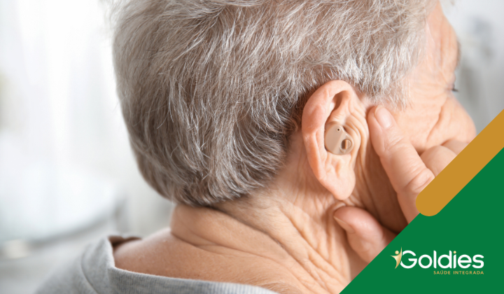 Idoso com cabelos grisalhos ajustando um aparelho auditivo no ouvido. A imagem inclui um logotipo que diz Goldies Saúde Integrada no canto inferior direito.