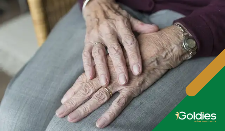 Mãos de idoso apoiadas no colo, com rugas visíveis, anéis e relógio no pulso. Vestem calças cinza. No canto inferior direito há um logotipo em verde e amarelo com o texto Goldies Saúde Integrada.