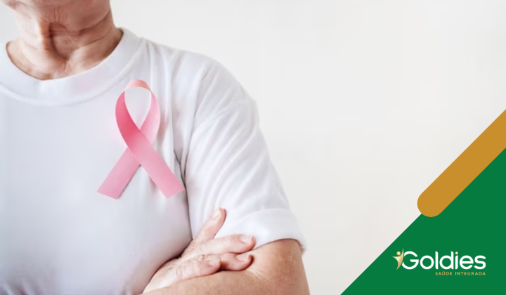 Uma pessoa vestindo uma camisa branca com uma fita rosa presa nela, simbolizando a conscientização sobre o câncer de mama, está de pé com os braços cruzados. O logotipo da Goldies Saúde Integrada é visível no canto inferior direito.