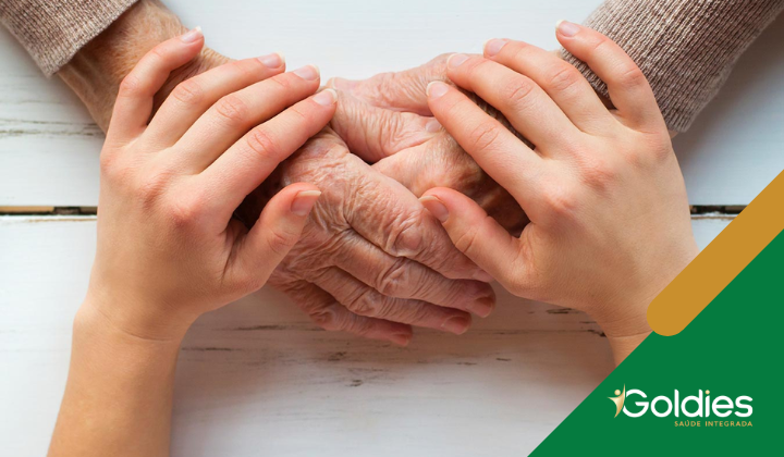 Dois pares de mãos apoiadas sobre uma superfície de madeira branca. As mãos mais velhas, com rugas, são gentilmente seguradas por mãos mais jovens. No canto inferior direito, há um logotipo verde e dourado com o texto Goldies Saúde Integrada.