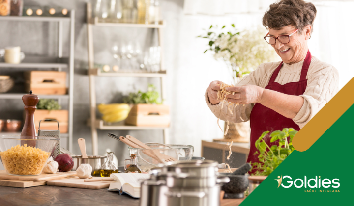 Uma mulher sorridente em uma cozinha segura macarrão. Vários ingredientes e utensílios de cozinha estão no balcão. Prateleiras ao fundo seguram pratos e potes. O logotipo Goldies Saúde Integrada está no canto.