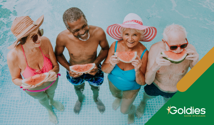Quatro adultos mais velhos, dois homens e duas mulheres, apreciam fatias de melancia em pé em uma piscina. Eles estão sorrindo e vestindo trajes de banho e chapéus. A imagem tem um logotipo Goldies no canto.