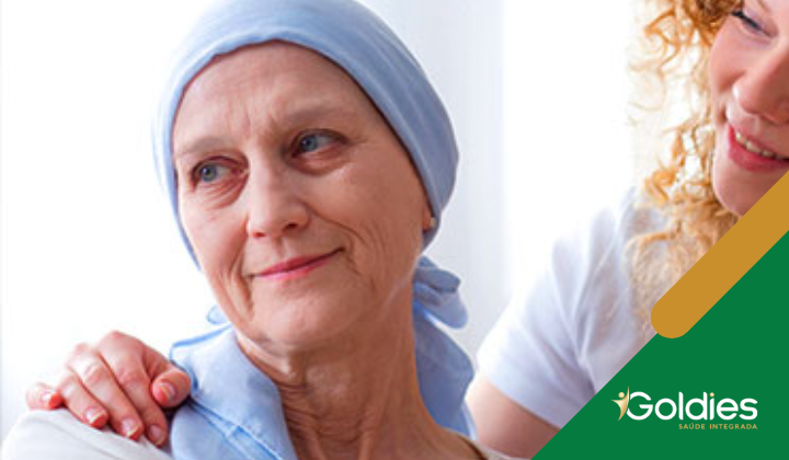 Uma mulher mais velha com um lenço na cabeça sorri gentilmente enquanto outra pessoa coloca uma mão reconfortante em seu ombro. A cena transmite apoio e cuidado. Um logotipo com a palavra Goldies é visível no canto inferior direito.