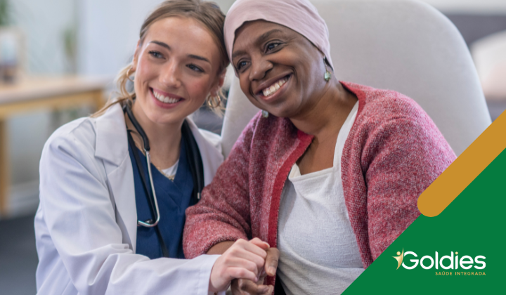 Um médico de jaleco branco e estetoscópio sorri enquanto segura as mãos de um paciente usando um lenço de cabeça e suéter rosa. Eles estão sentados em um ambiente de assistência médica. O logotipo Goldies Saúde Integrada está no canto inferior direito.