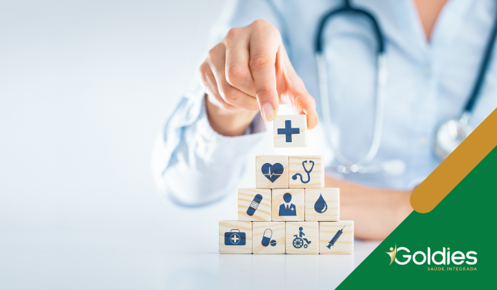 Uma pessoa empilhando blocos de madeira com ícones médicos, como um coração, estetoscópio, pílula e seringa. O bloco superior tem uma cruz médica. A pessoa usa um estetoscópio. O logotipo no canto diz Goldies Saúde Integrada.