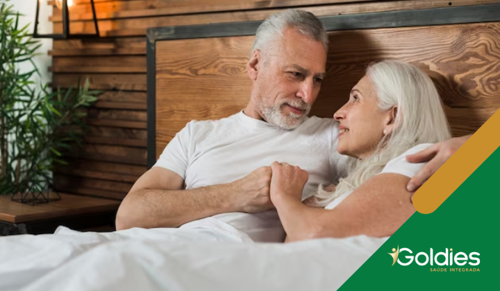 Um casal de idosos está deitado na cama, olhando um para o outro e sorrindo. Ambos estão vestindo camisas brancas. Há uma cabeceira de madeira e um vaso de planta ao fundo. As palavras Goldies Saúde Integrada são visíveis no canto inferior direito.