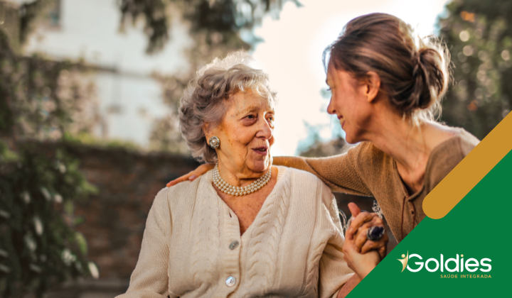 Uma mulher idosa com cabelo curto e grisalho está sentada do lado de fora, sorrindo e conversando com uma mulher mais jovem com cabelo preso para trás. Ambas estão usando tops de cor clara. A cena é calorosa e amigável. O logotipo Goldies Saúde Integrada é visível no canto.