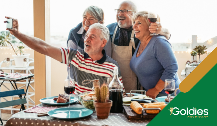 Quatro idosos sorrindo e tirando uma selfie em uma mesa de jantar ao ar livre com comida e bebidas. Eles parecem alegres e estão cercados por uma atmosfera calorosa e acolhedora. O logotipo Goldies Saúde Integrada é visível no canto.
