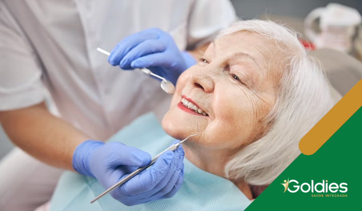 Uma mulher idosa com cabelos grisalhos sorri enquanto um profissional odontológico examina sua boca usando um espelho e uma ferramenta. Ela está usando um babador e está sentada em uma cadeira odontológica. O profissional está usando luvas azuis. A imagem tem o logotipo Goldies Saúde Integrada.