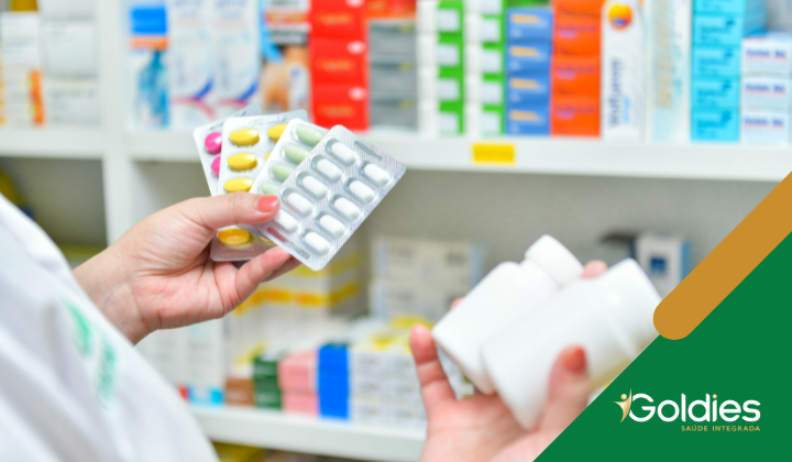Uma pessoa em uma farmácia segura vários blisters de comprimidos em uma mão e três frascos de comprimidos brancos na outra. Prateleiras estocadas com vários medicamentos são visíveis ao fundo. A marca Goldies Saúde Integrada é exibida no canto.