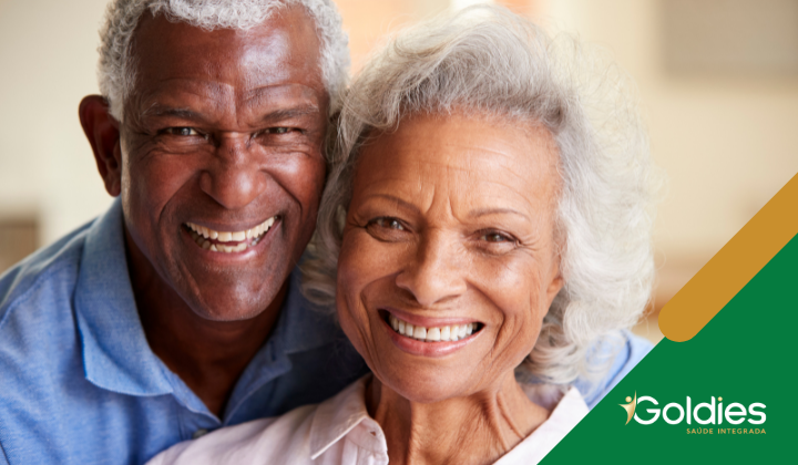 Homem e mulher idosos sorrindo calorosamente para a câmera. Eles estão próximos, parecendo felizes. A imagem tem um design de canto verde e dourado com o texto Goldies Saúde Integrada no canto inferior direito.