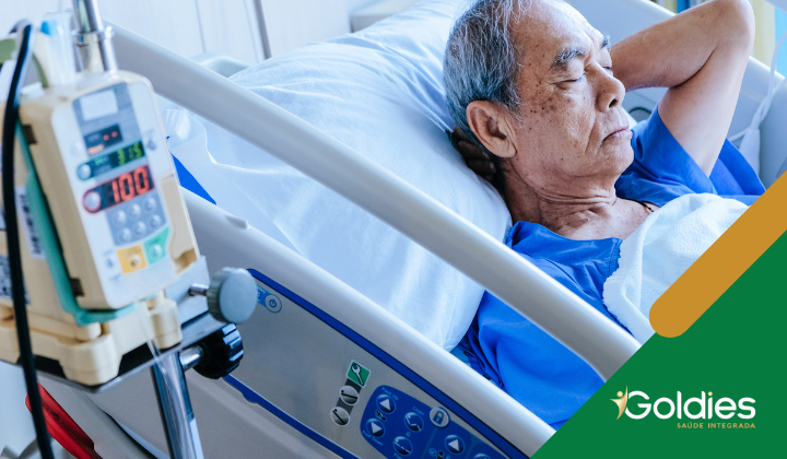 Homem idoso em uma cama de hospital, descansando com os olhos fechados. Equipamento médico é visível ao lado dele. A imagem apresenta um logotipo no canto com o texto Goldies Saude Integrada.