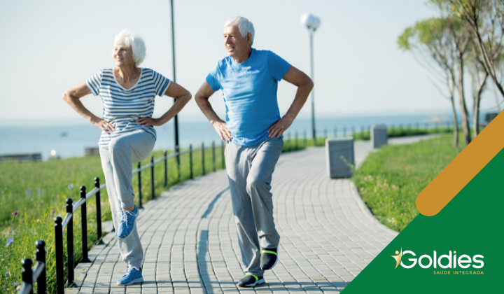 Dois idosos se exercitando em um caminho pavimentado perto do mar, vestindo roupas esportivas casuais. Eles estão realizando um levantamento de pernas, parecendo alegres e cheios de energia. A cena é ensolarada, com grama verde e um céu azul claro. Logotipo Goldies no canto.