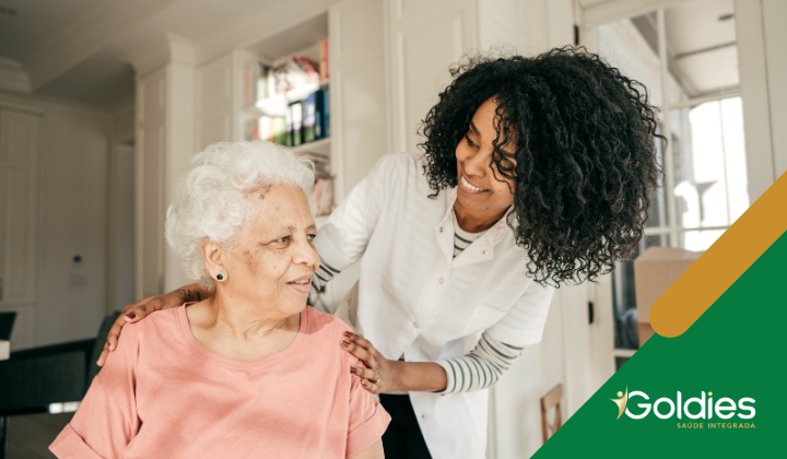 Uma cuidadora, sorrindo, se inclina sobre uma senhora idosa de cabelos brancos, que está sentada e olhando para cima. Elas estão em uma sala iluminada. O logotipo Goldies Saúde Integrada está no canto inferior direito.