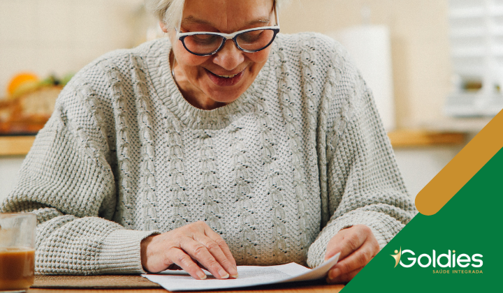 Uma pessoa idosa de óculos sorri enquanto lê um documento em uma mesa. Ela está usando um suéter de tricô. Uma xícara de café está próxima. A imagem tem o logotipo da Goldies Saúde Integrada no canto.