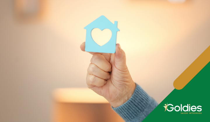 Uma mão segurando um pequeno recorte de casa azul com um buraco em forma de coração no centro ilustrando a postagem sobre como tornar a casa segura e confortável com 5 dicas de adaptação para seniores