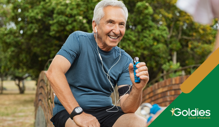 Homem idoso em traje esportivo sorrindo enquanto está sentado em um banco em um parque. Ele segura um inalador em uma mão e usa fones de ouvido. Árvores verdes exuberantes e grama são visíveis no fundo, com um design de canto de marca na imagem.