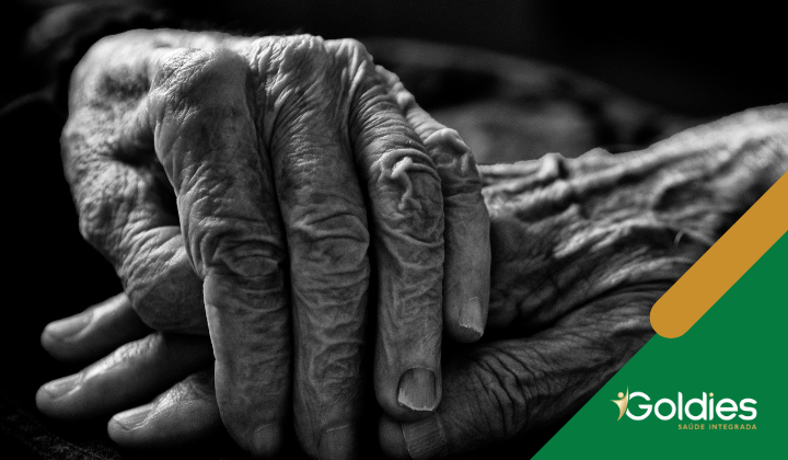 Close de duas mãos idosas gentilmente entrelaçadas em uma imagem em preto e branco. As mãos são detalhadas e enrugadas, simbolizando idade e experiência. O logotipo no canto diz Goldies Saúde Integrada.