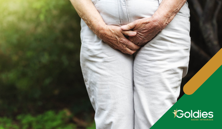 Pessoa idosa em pé ao ar livre com as mãos entrelaçadas na frente, vestindo calças de cor clara. O fundo é folhagem verde. Um logotipo verde e dourado com o texto Goldies está no canto inferior direito.