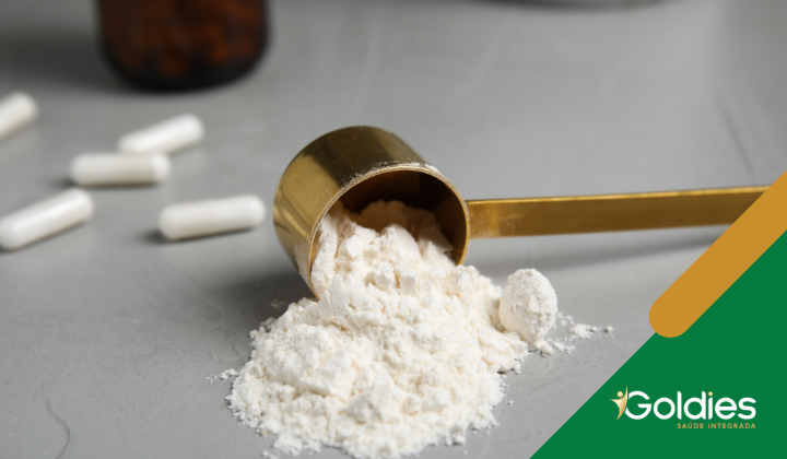 Uma colher dourada com pó branco é derramada sobre uma superfície cinza. Várias cápsulas brancas estão espalhadas por perto. O logotipo da Goldies Saúde Integrada está no canto.