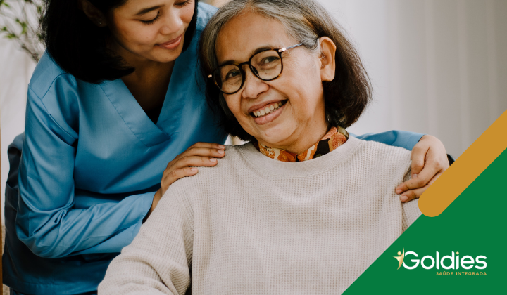Uma senhora idosa sorridente usando óculos e um suéter leve está sentada enquanto uma mulher mais jovem em uniforme azul gentilmente apoia as mãos em seus ombros. O logotipo Goldies Saúde Integrada é visível no canto inferior.
