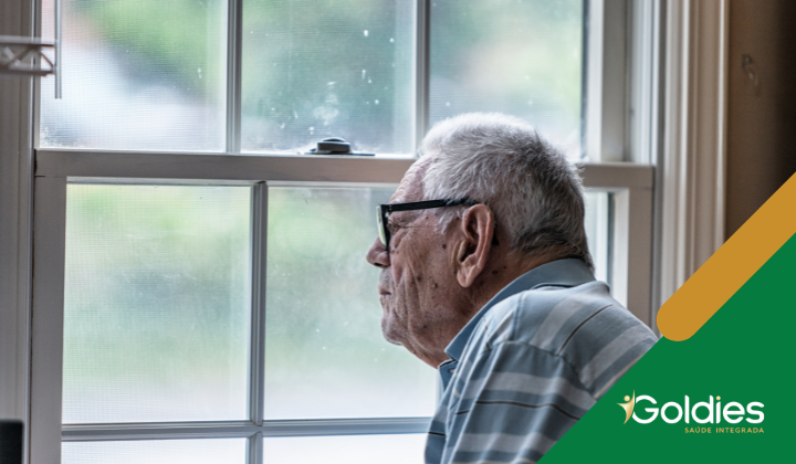 Homem idoso usando óculos e camisa listrada olha pela janela, com árvores visíveis através do vidro. Há um logotipo Goldies Saúde Integrada no canto.