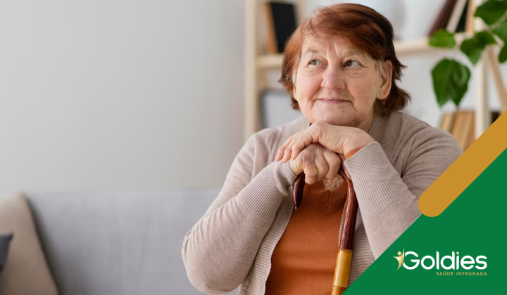 Uma mulher idosa com cabelo curto e um suéter quente está sentada em um sofá, segurando uma bengala, olhando pensativamente para o lado. Prateleiras com livros e uma planta estão ao fundo. O logotipo Goldies está no canto inferior direito.