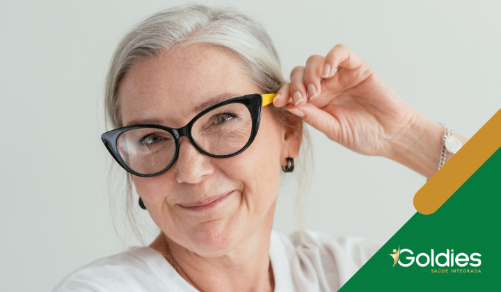 Uma mulher mais velha com cabelos grisalhos sorri enquanto ajusta seus óculos de aro preto. Ela usa uma camisa branca e brincos. Um logotipo com Goldies está no canto inferior direito.