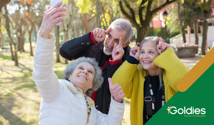 Três amigos idosos tiram uma selfie alegremente ao ar livre em um parque. Eles estão sorrindo e fazendo gestos brincalhões com os dedos na cabeça. O cenário é ensolarado com árvores e vegetação ao fundo. O canto inferior direito tem Goldies Saúde Integrada escrito nele.