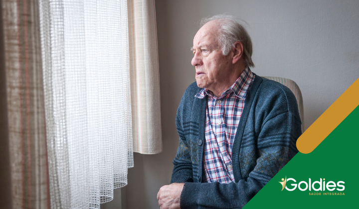 Um homem idoso com uma camisa xadrez e um cardigan está sentado perto de uma janela, parecendo contemplativo. O quarto é suavemente iluminado, com cortinas transparentes. O logotipo Goldies é visível no canto.