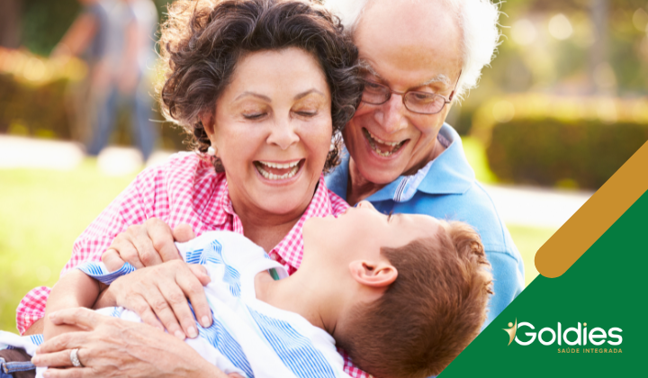 Casal de idosos interagindo alegremente com um menino em um parque. A mulher veste uma camisa xadrez rosa, o homem tem uma camisa azul e o menino está deitado no colo deles, sorrindo. O logotipo Goldies Saúde Integrada está no canto.