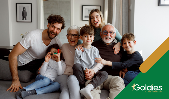 Uma família feliz multigeracional posa junta em uma sala de estar. Dois avós sentam-se no centro com seus netos. Dois adultos, um homem e uma mulher, estão nas laterais. Há um logotipo Goldies Saúde Integrada no canto inferior direito.