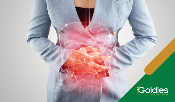 Uma pessoa de blazer azul segurando o estômago, com uma ilustração digital do sistema digestivo brilhando em vermelho sobrepondo o tronco, indicando desconforto. O canto inferior direito apresenta o logotipo da Goldies Saúde Integrada.