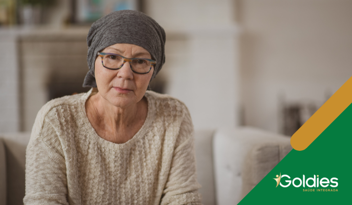Idoso usando óculos e lenço na cabeça sentado em um sofá, olhando para a câmera. Eles vestem um suéter bege. Um desenho verde e laranja com o texto Goldies Saúde Integrada está no canto inferior direito.