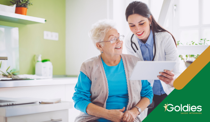 Um profissional de saúde sorridente mostra um tablet para uma senhora idosa em uma sala aconchegante e iluminada. A mulher está sentada e vestindo uma blusa azul-clara e um colete bege. A profissional veste um jaleco branco e estetoscópio. O logotipo da Goldies Saúde Integrada é visível.