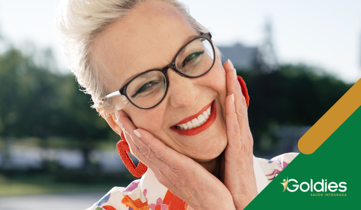 Pessoa sorridente usando óculos e brincos vermelhos brilhantes segura o rosto com as duas mãos. Ela está ao ar livre, vestindo uma roupa colorida. O canto inferior tem o logotipo Goldies Saúde Integrada.